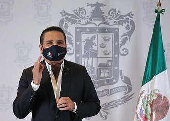 Muñeco con máscara y traje formal frente a escudo y bandera de México.