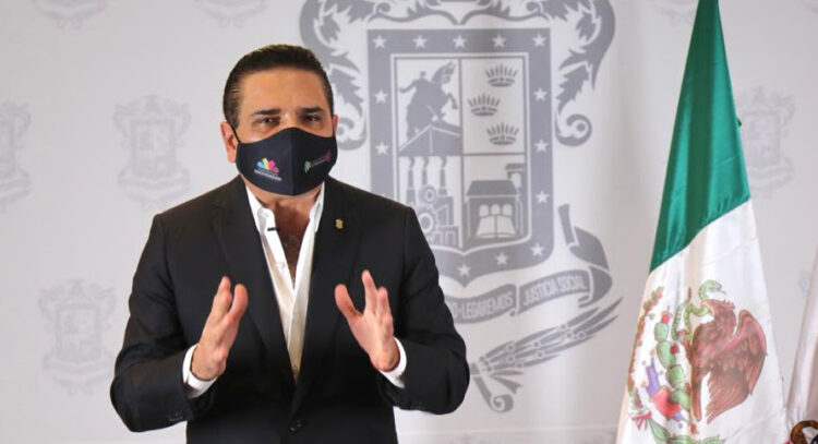 Prominente figura política mexicana con mascarilla y fondo oficial con bandera de México.