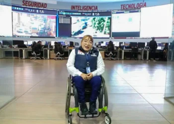 Personaje en silla de ruedas en centro de control de seguridad con pantallas de tecnología avanzada en fondo.